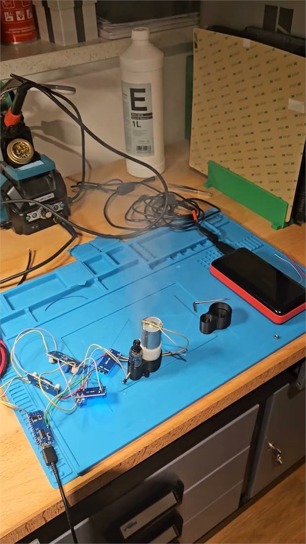 Workbench with a blue soldering mat, microcontroller boards, wiring and a small fog module under test.